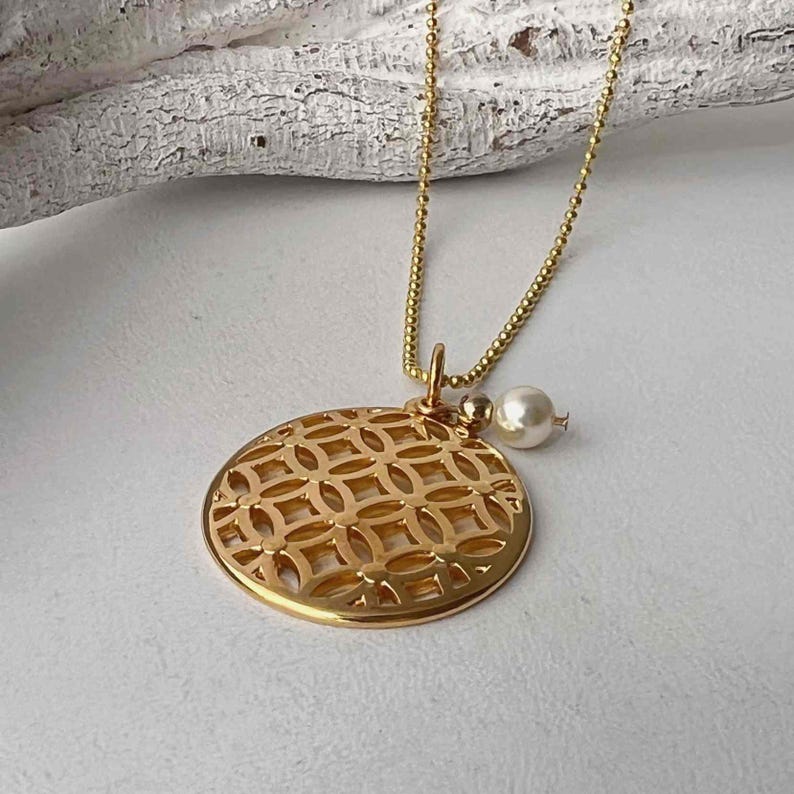 Puede incluir: Un collar dorado con un colgante redondo calado con un patr&oacute;n geom&eacute;trico repetitivo. Un peque&ntilde;o dije de perla cuelga de la cadena. El collar se muestra sobre una superficie blanca.
