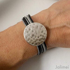 K&ouml;nnte beinhalten: Ein mehrreihiges Lederarmband mit einem geh&auml;mmerten, silberfarbenen runden Anh&auml;nger. Das Armband besteht aus schwarzem und grauem Leder.