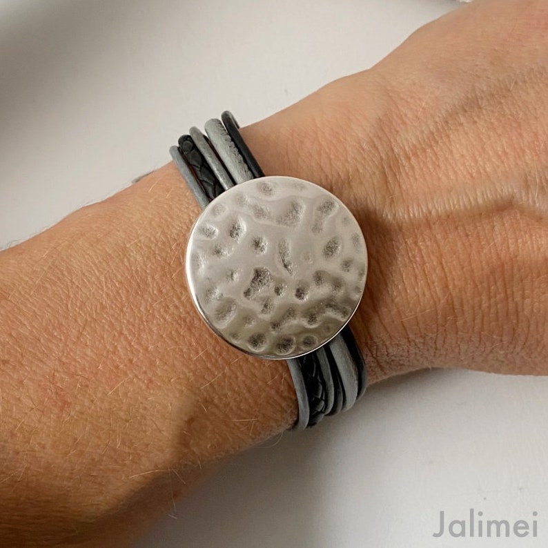 K&ouml;nnte beinhalten: Ein mehrreihiges Lederarmband mit einem geh&auml;mmerten, silberfarbenen runden Anh&auml;nger. Das Armband besteht aus schwarzem und grauem Leder.