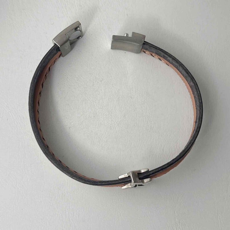 May include: A brown leather bracelet with silver metal clasps. The bracelet is made of two pieces of leather that are stitched together.