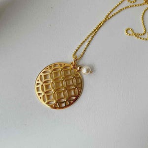 Puede incluir: Collar dorado con colgante circular calado con dise&ntilde;o geom&eacute;trico. Un peque&ntilde;o dije de perla cuelga del colgante. La cadena del collar es de estilo fino de bolitas.