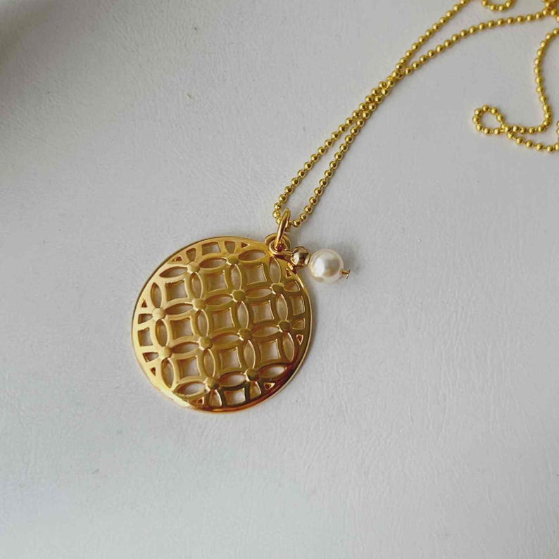 Puede incluir: Collar dorado con colgante circular calado con dise&ntilde;o geom&eacute;trico. Un peque&ntilde;o dije de perla cuelga del colgante. La cadena del collar es de estilo fino de bolitas.