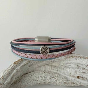 May include: A multi-strand bracelet with pink, blue, and black leather cords. The bracelet has a silver magnetic clasp and a spiral charm. The braided leather adds texture to the design.