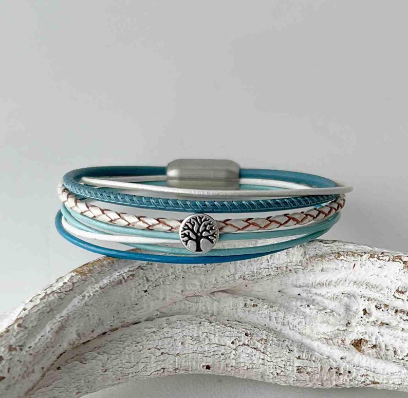 May include: A multi-layered bracelet with a silver tree of life charm. The bracelet features shades of blue and white leather straps.