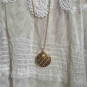 Puede incluir: Un collar dorado con un colgante redondo con un patr&oacute;n geom&eacute;trico y una peque&ntilde;a perla blanca en el centro. El collar cuelga sobre un fondo de tela blanca y con encaje.