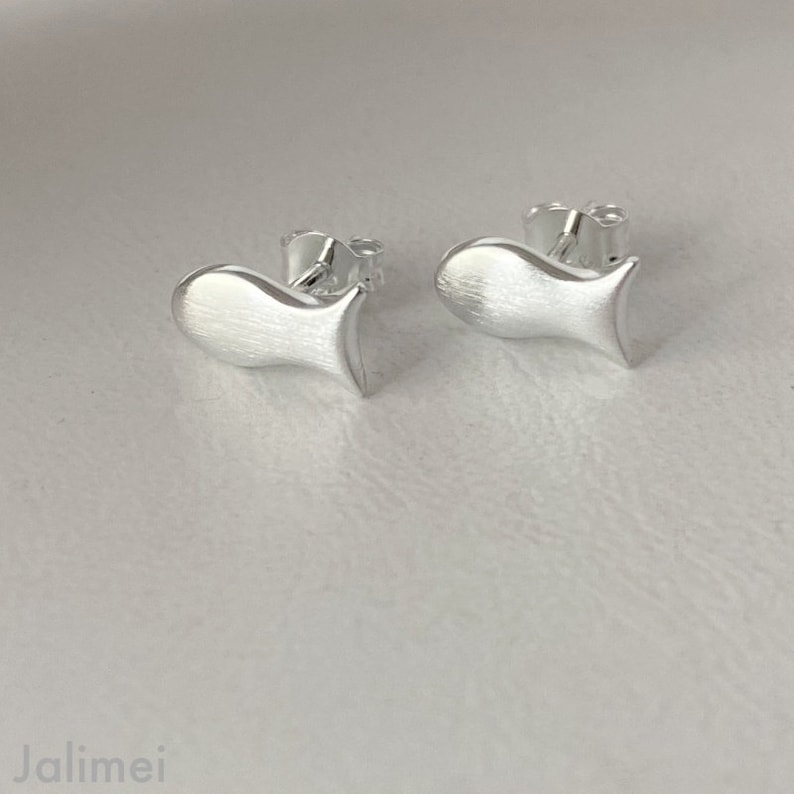 K&ouml;nnte beinhalten: Ein Paar silberne Ohrstecker in Fischform. Die Ohrringe sind klein und zart, mit einer geb&uuml;rsteten Oberfl&auml;che.