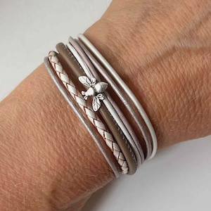 Könnte beinhalten: Ein mehrlagiges Armband mit einem geflochtenen Lederband und mehreren glatten Lederbändern in Braun- und Weißtönen. Ein kleiner silberner Bienenanhänger ist am Armband befestigt.