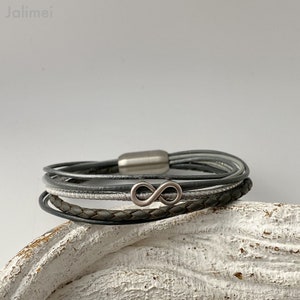 Puede incluir: Un brazalete de cuero multicapa con un dije de plata en forma de infinito. El brazalete est&aacute; hecho de cuero gris y blanco y tiene un cierre magn&eacute;tico.