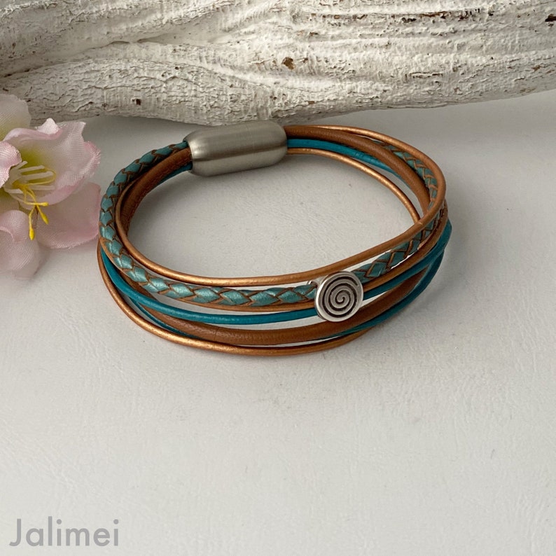 May include: A multi-strand leather bracelet with a braided turquoise and copper leather design. The bracelet has a silver magnetic clasp and a silver swirl charm.