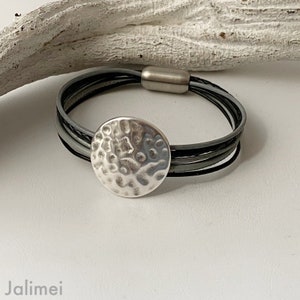 K&ouml;nnte beinhalten: Ein silbernes Armband mit einem geh&auml;mmerten Metall-Scheibenanh&auml;nger. Das Armband besteht aus mehreren Str&auml;ngen grauen Leders.