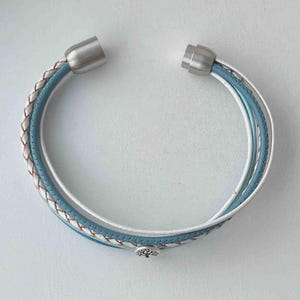 May include: A multi-layered bracelet with a blue, white, and brown braided leather design. The bracelet has silver magnetic clasps and a silver tree charm.