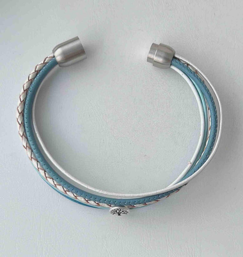 May include: A multi-layered bracelet with a blue, white, and brown braided leather design. The bracelet has silver magnetic clasps and a silver tree charm.