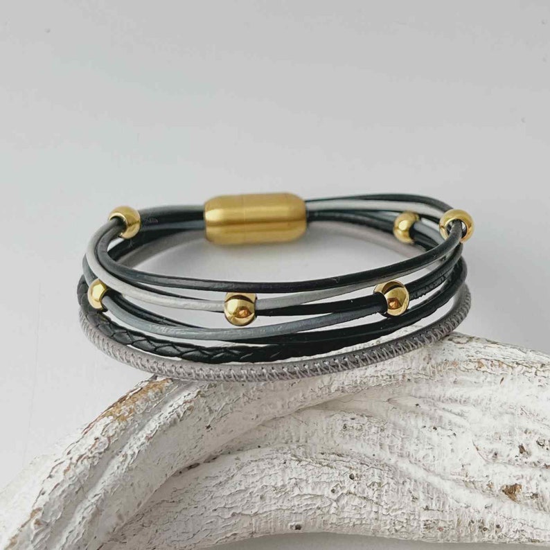 K&ouml;nnte beinhalten: Ein mehrreihiges Lederarmband mit einem Magnetverschluss. Das Armband besteht aus schwarzen, grauen und wei&szlig;en Lederstr&auml;ngen mit goldfarbenen Perlen.