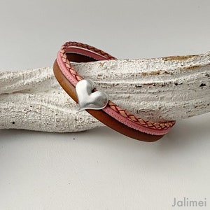 Pode incluir: Um bracelete de couro castanho com uma trança de couro rosa e um fecho de coração prateado.