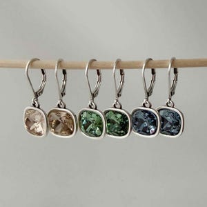 Può includere: Sono mostrati sei paia di orecchini pendenti quadrati con chiusura a leva. Gli orecchini hanno montature color argento e sono disponibili nei colori champagne, marrone, verde e blu. Gli orecchini sono esposti su un'asta di legno.