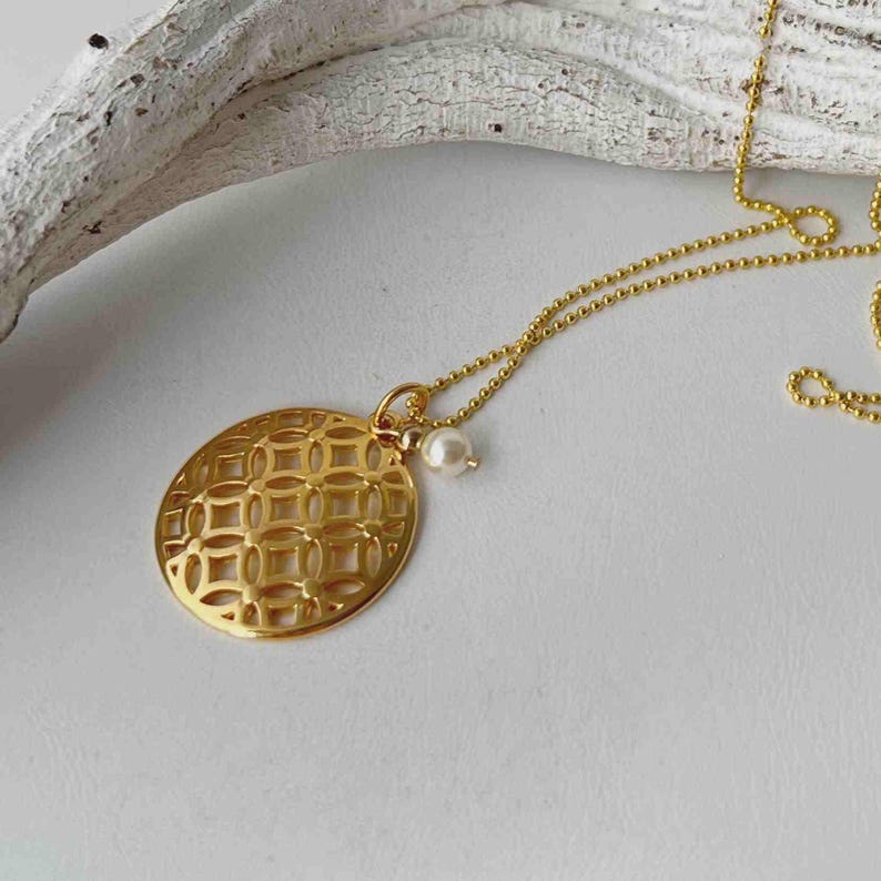 Puede incluir: Un collar con colgante dorado con un dise&ntilde;o circular calado. El colgante cuelga de una fina cadena y est&aacute; adornado con una peque&ntilde;a perla blanca. El collar se presenta sobre una superficie blanca.