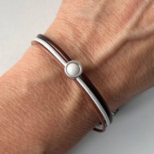 Könnte beinhalten: Ein Lederarmband mit einem braunen und weißen Band und einem silberfarbenen Verschluss mit einem weißen kreisförmigen Stein. Das Armband wird am Handgelenk getragen.