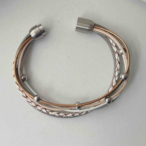 May include: A multi-strand bracelet with a braided design in white, grey, and brown leather. The bracelet has silver magnetic clasps and silver beads.