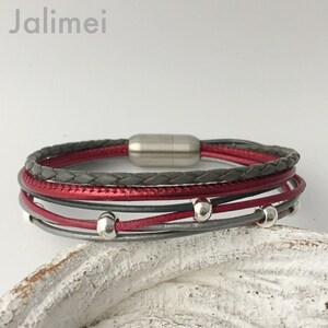 May include: A multi-layered leather bracelet with a magnetic clasp. The bracelet features a braided grey leather strap, a red leather strap, and a silver bead accent.