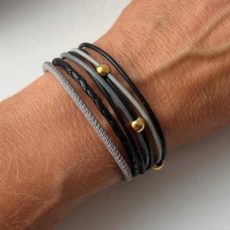 K&ouml;nnte beinhalten: Ein Stapel Lederarmb&auml;nder in Schwarz und Grau mit goldenen Akzenten. Die Armb&auml;nder sind &uuml;bereinander am Handgelenk einer Person gelegt.