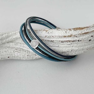 Könnte beinhalten: Ein mehrsträngiges Lederarmband mit einem silbernen Spiral-Anhänger. Das Armband besteht aus blauen, schwarzen und grauen Lederbändern.