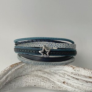 K&ouml;nnte beinhalten: Ein mehrlagiges Armband mit einem silbernen Sternanh&auml;nger. Das Armband besteht aus blauem, wei&szlig;em und schwarzem Leder und Stoff.