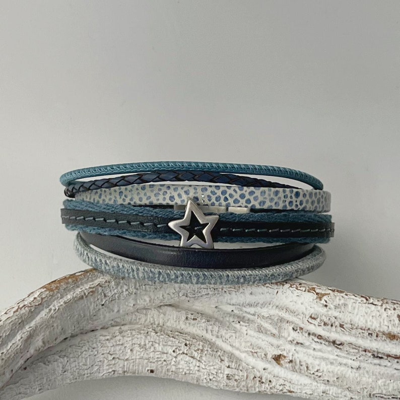 K&ouml;nnte beinhalten: Ein mehrlagiges Armband mit einem silbernen Sternanh&auml;nger. Das Armband besteht aus blauem, wei&szlig;em und schwarzem Leder und Stoff.