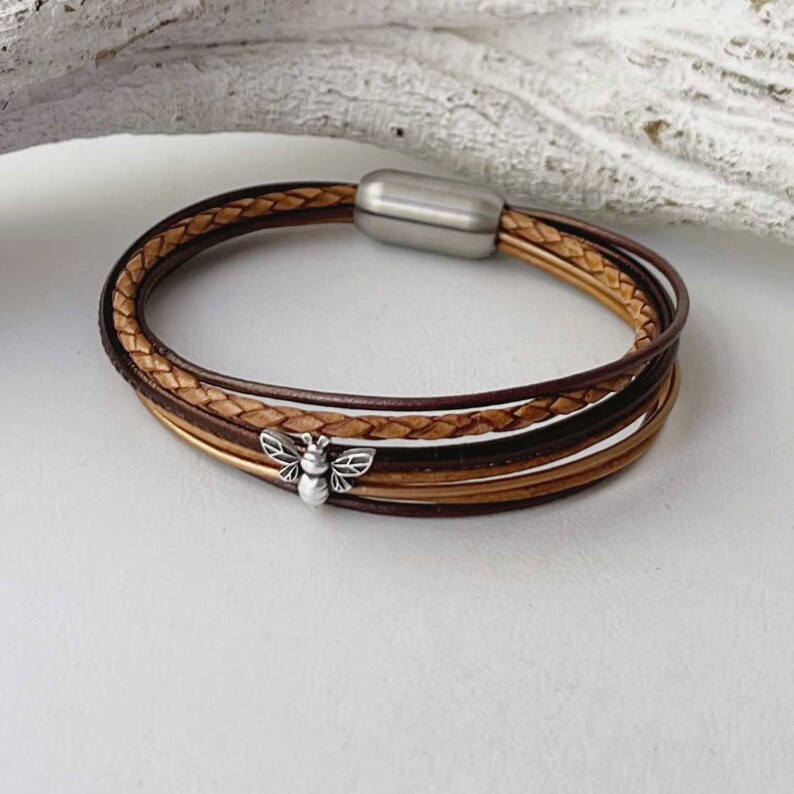 May include: A multi-strand leather bracelet with a silver bee charm. The bracelet is made of brown and tan leather and has a magnetic clasp.