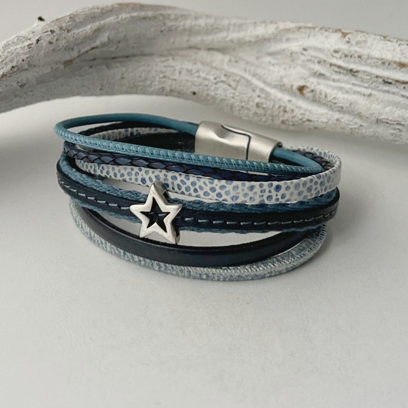 K&ouml;nnte beinhalten: Ein mehrlagiges Armband mit einem silbernen Sternanh&auml;nger. Das Armband besteht aus blauem und schwarzem Leder und hat einen magnetischen Verschluss.