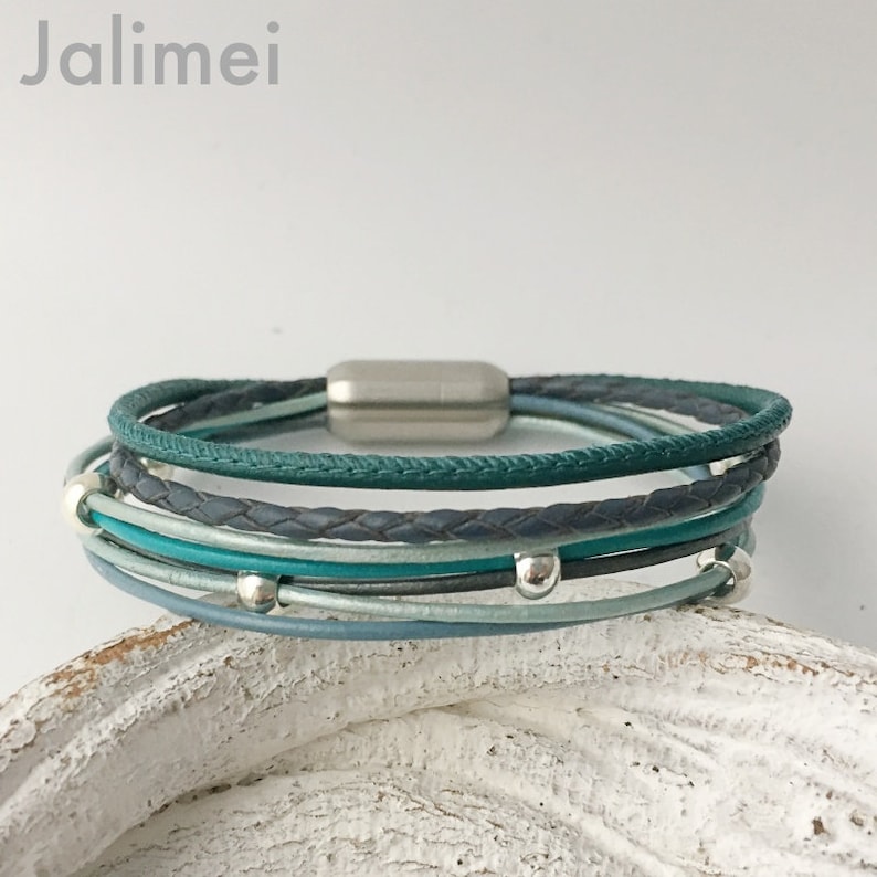 May include: Multi-layered leather bracelet with a teal, gray, and blue color scheme. The bracelet has a silver magnetic clasp and silver beads.