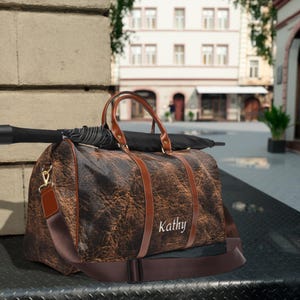 Könnte beinhalten: Eine braune Leder-Reisetasche mit einem personalisierten Namen "Kathy", der auf der Vorderseite gestickt ist. Die Tasche hat einen braunen Lederriemen und ein schwarzer Regenschirm liegt auf der Tasche.