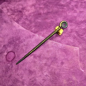 May include: A twisted metal hair stick with a decorative loop at the top. The stick is dark brown and has a rustic finish.