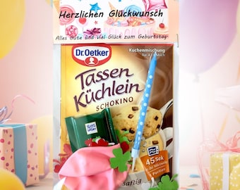 May include: A birthday gift set with a box of Dr. Oetker Tassen Küchlein Schokino chocolate cake mix, a pink balloon, a blue candle, and a small box of Rücker Sport chocolate. The packaging has the text "Herzlichen Glückwunsch" and "Alles Gute und viel Glück zum Geburtstag".