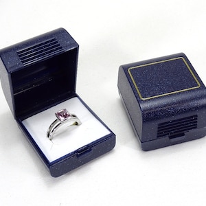 Könnte beinhalten: Ein silberner Ring mit einem quadratischen, hellvioletten Edelstein liegt in einer offenen, marineblauen Ringschatulle mit weißem Innenraum. Eine geschlossene, passende Schachtel befindet sich daneben. Die Schachteln haben einen goldenen Rand.