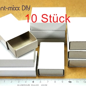 May include: Ten white cardboard matchboxes stacked in two piles. The boxes are blank and have a sliding drawer. The text "bunt-mixx DIY 10 Stück" is visible in the image.