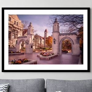 May include: A framed landscape photograph depicting the Sample Gates at Indiana University. The image features stone archways, a clock tower in the distance, and autumn foliage under a clear sky, all enclosed within a simple black frame.