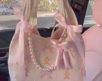 May include: A pink knitted tote bag with a gingerbread man pattern, pink ribbon handles, and a pearl necklace. The bag has a pink bow charm and a pink beaded bow charm.