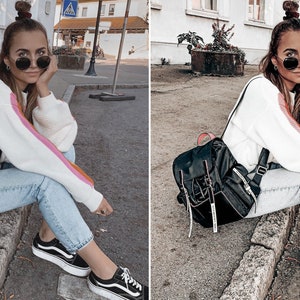 Peut inclure: Une femme portant un pull blanc &agrave; rayures roses et orange, un jean bleu et des baskets noires. Elle porte un sac &agrave; dos noir avec des bretelles blanches. Elle est assise sur un trottoir devant un b&acirc;timent.