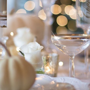 May include: A white table setting with a champagne flute, white pumpkins, and white roses. The table is decorated with a white tablecloth and gold accents.