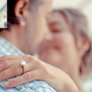 May include: A close-up of a hand wearing a silver ring with a large, sparkling diamond. The ring is on the finger of a person wearing a blue and white plaid shirt. The person's face is out of focus in the background.