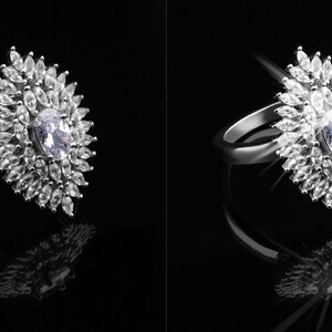 May include: A silver ring with a large, oval-shaped diamond surrounded by smaller diamonds. The ring is set against a black background and is reflecting light.