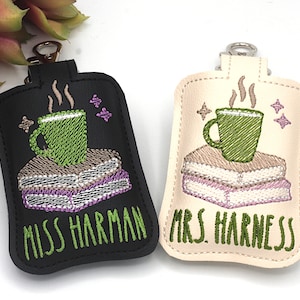 May include: Two keychains with embroidered designs. One is black, the other is cream-colored. Each features a green mug atop a stack of books, with steam rising. The black keychain reads "MISS HARMAN" in green, while the cream one says "MRS. HARNESS."