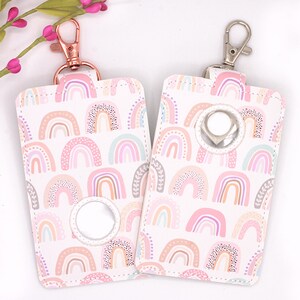 May include: Two white, rectangular dog waste bag holders with a pastel rainbow pattern. Each holder has a metal clip and a circular opening for bag dispensing. One clip is rose gold, the other is silver.