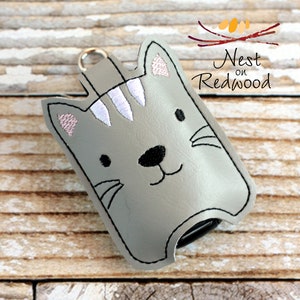 May include: A gray cat-shaped keychain holder with a black outline, featuring embroidered details. The cat has a black nose, whiskers, and a smiling mouth. The ears have pink embroidered accents. The text "Nest on Redwood" is in the upper right corner.