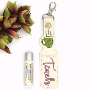 May include: A cream-colored keychain with a green coffee cup design and the embroidered word "Teach" in purple. A lip balm and a succulent plant are also in the image. The keychain has a silver clasp.