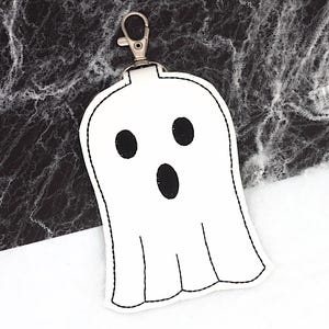 May include: A white ghost-shaped keychain with a black outline and three black oval-shaped facial features. The keychain has a silver-colored clasp at the top. The background is a mix of black and white.