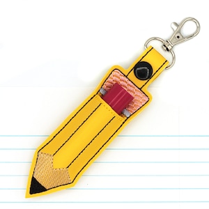 May include: A yellow pencil-shaped keychain with black stitching and a silver clasp. The pencil case holds a red eraser. The pencil tip is black and tan. The background is a white surface with blue lines.