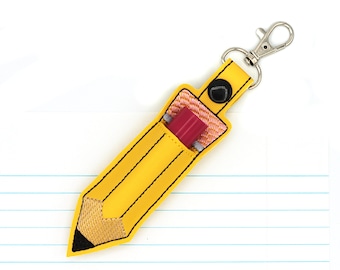 May include: A yellow pencil-shaped keychain with black stitching and a silver clasp. The pencil case holds a red eraser. The pencil tip is black and tan. The background is a white surface with blue lines.
