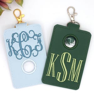 May include: Two personalized keychains. One is light blue with a teal monogram, the other is dark green with a yellow monogram. Both have a gold or silver clasp and a circular opening.