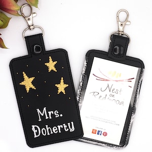 May include: Two black rectangular ID badge holders with silver swivel lobster clasps. One holder features embroidered gold stars and the white text Mrs. Doherty. The other holder has a clear plastic window displaying a card with the text Nest on Redwood.